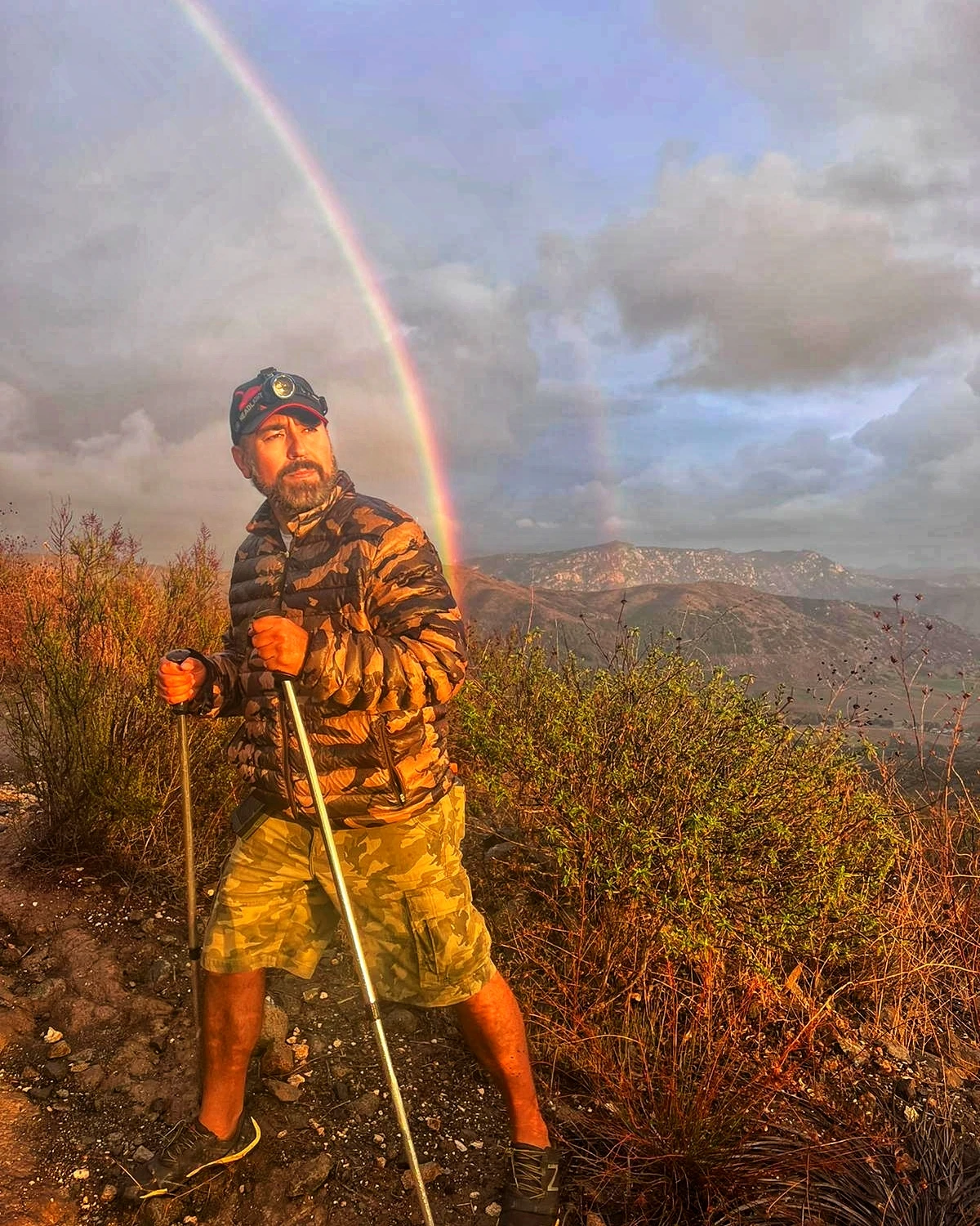 A hiker standing beneath a rainbow in mountain weather
